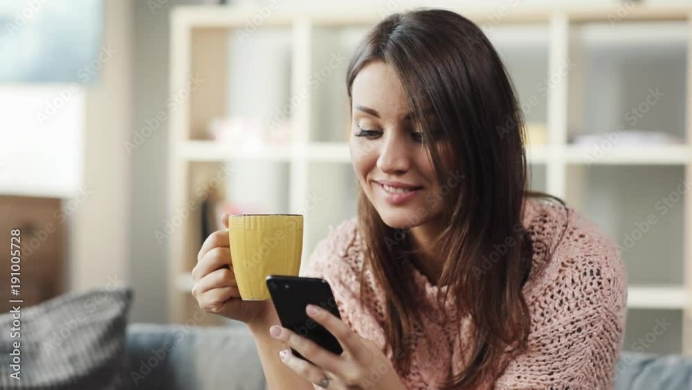 Pretty young brown-haired woman in pink sweater sitting on sofa in living room, looking at smartphone's screen, writing message. Cute girl looking dreamily out the window Pretty young brown-haired woman in pink sweater sitting on sofa in living room, looking at smartphone's screen, writing message. Cute girl looking dreamily out the window