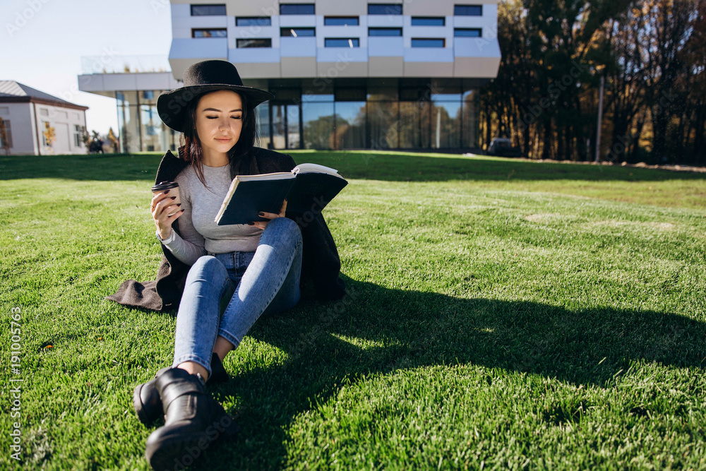 Fototapeta premium Stylish girl in black hat and coat sits with book and coffee on the lawn before the library