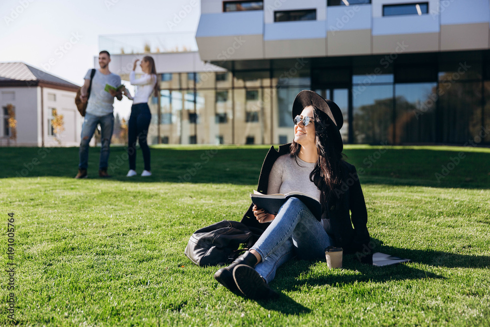 Stylish girl in black hat and coat sits with book and coffee on the lawn before the library