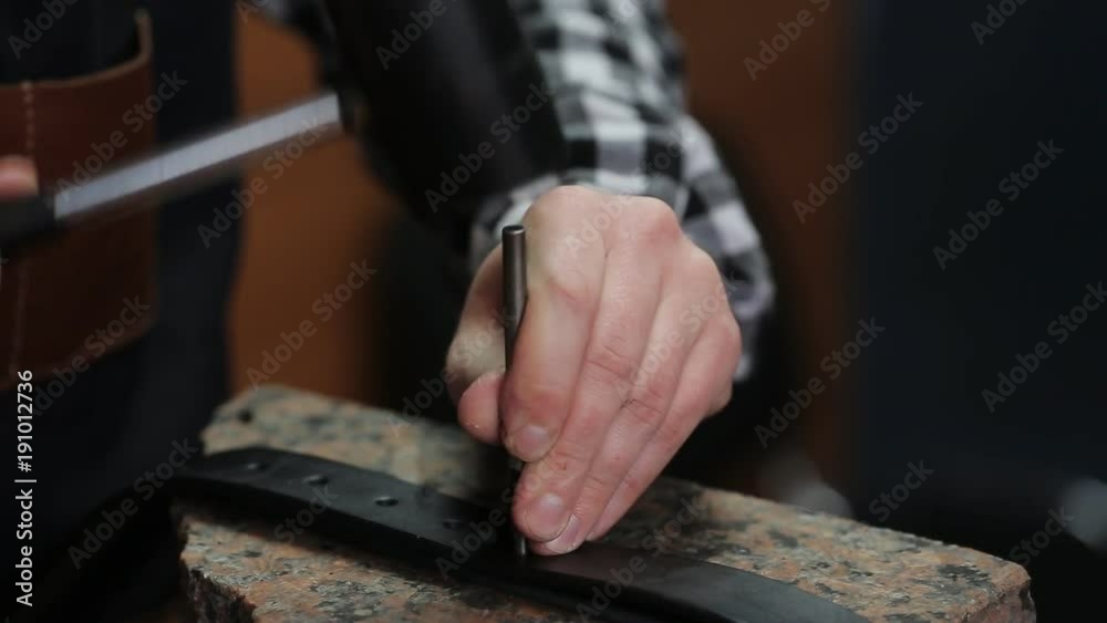 Working process of the leather belt in the leather workshop. Man holding crafting tool and working. He is placing billet to sturdy stand, fixing round knife on a mark and hitting with a small hammer Working process of the leather belt in the leather workshop. Man holding crafting tool and working. He is placing billet to sturdy stand, fixing round knife on a mark and hitting with a small hammer