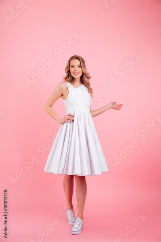 Full-length photo of charming blonde woman looking on camera with smile and holding copyspace on her palm, isolated over pink background