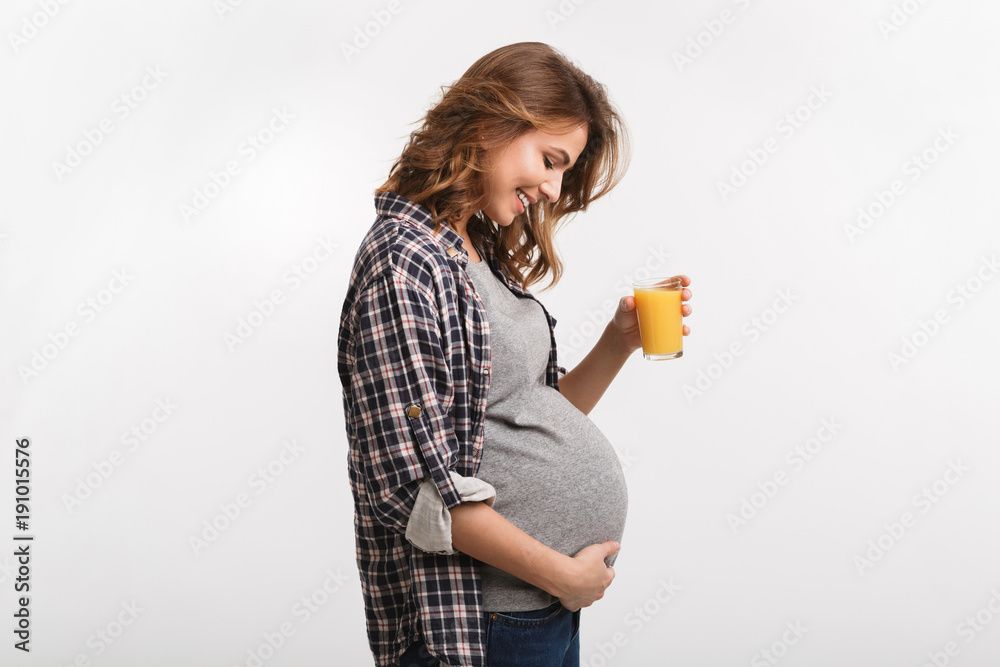 side view of smiling pregnant woman holding glass with juice and touching belly isolated on white