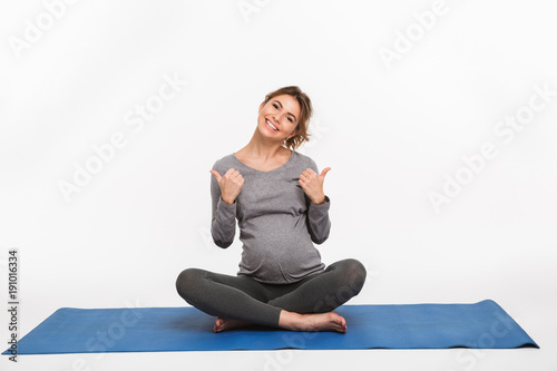happy pregnant woman sitting on yoga mat and showing thumbs up isolated on white