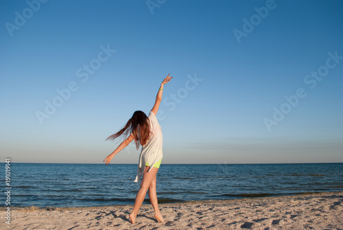 young beautiful girl dances on the seashore, her long hair is waving, she stands on the sand against the blue sea