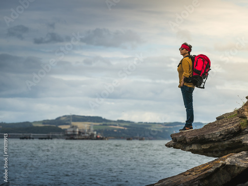 A tourist woman of  middle age with a backpack, standing on the rocks and looking at the horizon.