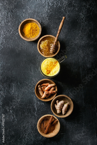 Glass of ayurvedic drink golden milk turmeric latte with curcuma powder and ingredients in wooden bowls above over black texture background. Top view, space