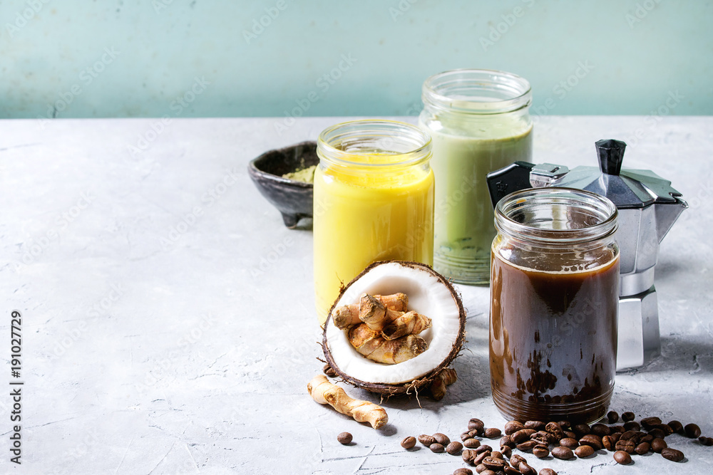 Variety of iced colorful latte drinks. Iced coffee, turmeric and matcha latte cocktails in glass jars with ingredients above over grey green texture background. Copy space Variety of iced colorful latte drinks. Iced coffee, turmeric and matcha latte cocktails in glass jars with ingredients above over grey green texture background. Copy space