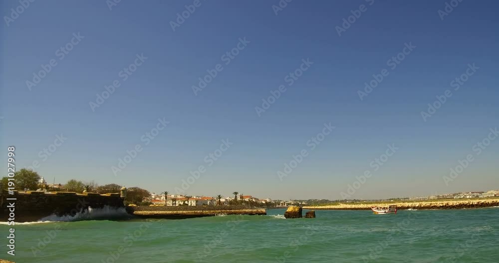 Waves crashing on the wall of fort Ponta da Bandeira in the background, and a boat with tourists entering the marina, Lagos Algarve Waves crashing on the wall of fort Ponta da Bandeira in the background, and a boat with tourists entering the marina, Lagos Algarve