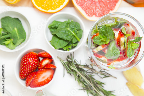 A glass of detox water with sliced strawberry and mint, cups with ingredients and a spring of rosemary, white background, top view