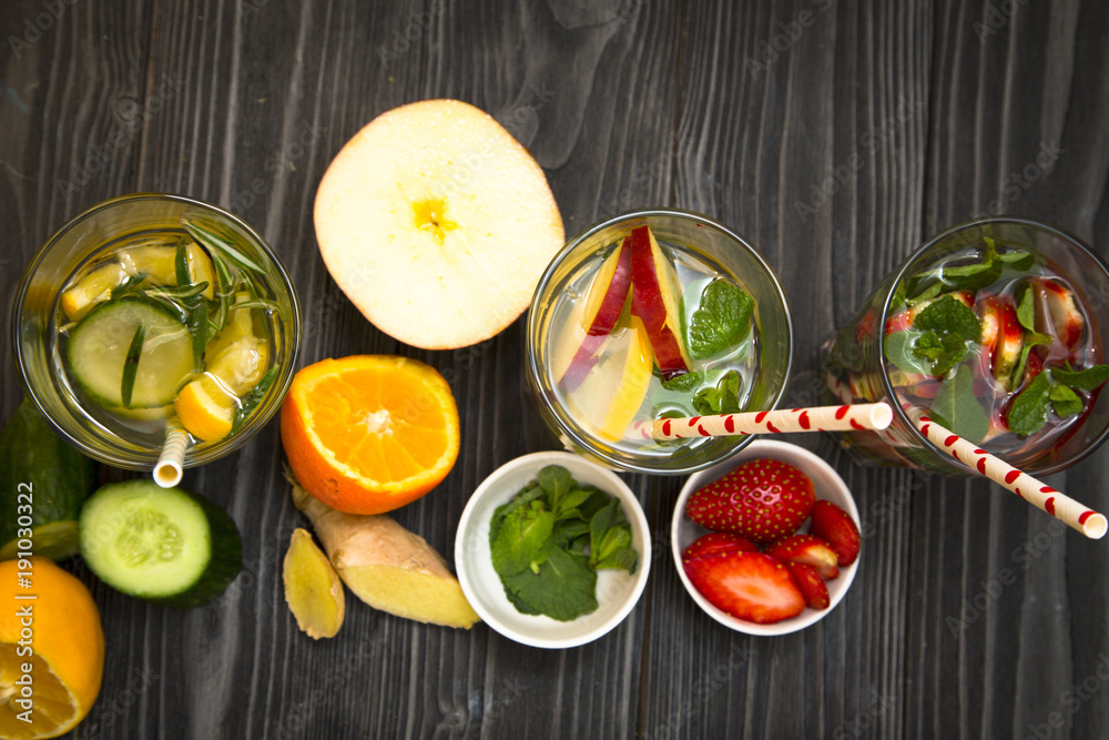 Glasses of detox water with sliced fruit, vegetables, berries and herbs, set of ingredients: banana, strawberry, orange, ginger, lemon, cucumber and herbs on black wood top view Glasses of detox water with sliced fruit, vegetables, berries and herbs, set of ingredients: banana, strawberry, orange, ginger, lemon, cucumber and herbs on black wood top view