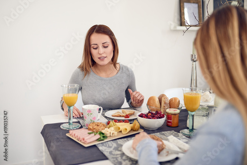 Smiling woman having breakfast with her daughter or friend sitting at the table with a healthy spread of fruit, cereal, bread rolls, cold meats, cheese and fresh orange juice