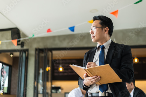 Asian businessman holding a book and pen to write a business plan. He standing and looking forward, thinking for business future and financial success.