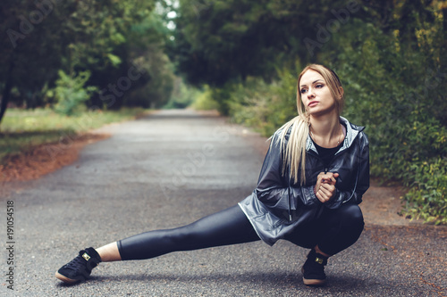 Attractive young woman doing stretching sports exercises in a city park at rainy weather.