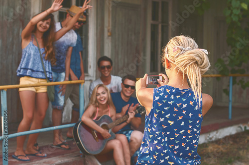 Young blonde girl with dreadlocks hairstyle takes a photo of a groups of her friends with her smartphone near the wooden holiday cabin
