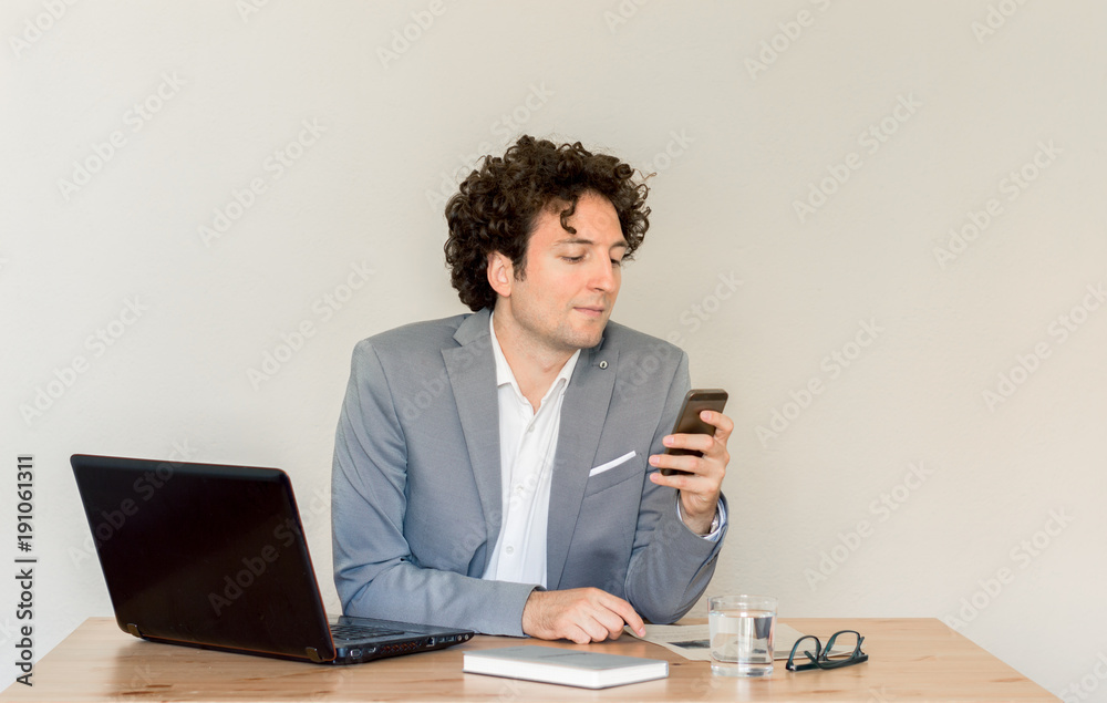Young Caucasian businessman sitting at his desk and using his mobile phone in front of empty clear wall concept Young Caucasian businessman sitting at his desk and using his mobile phone in front of empty clear wall concept