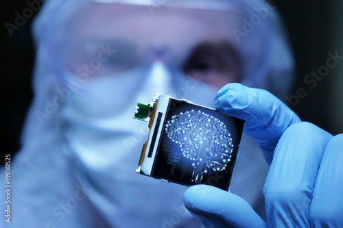 An Ultra Modern Electronic Manufacturing Factory Design Engineer in Sterile Coverall Holds Microchip with symbols in futuristic holography