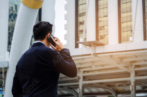 back view of young smart business man wearing black suit making phone call with mobile smart phone in building city background, internet network connection, technology communication, financial concept