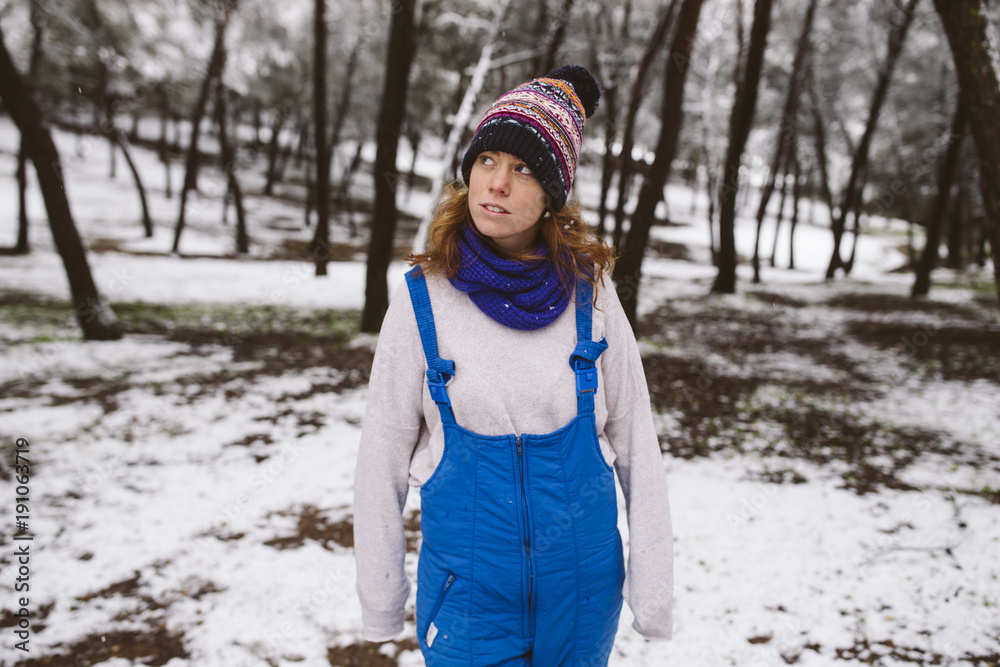 Redhead woman smiling in the snow. She wears a blue overall, a white sweater, purple scarf and a colorful hat.