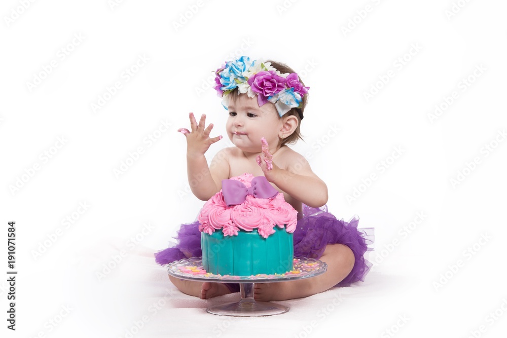 Cute happy baby girl with blue eyes is tasting the cake and purple butter icing on her sticky fingers from her first birthday cake smash while sitting on a white background . Cute happy baby girl with blue eyes is tasting the cake and purple butter icing on her sticky fingers from her first birthday cake smash while sitting on a white background .