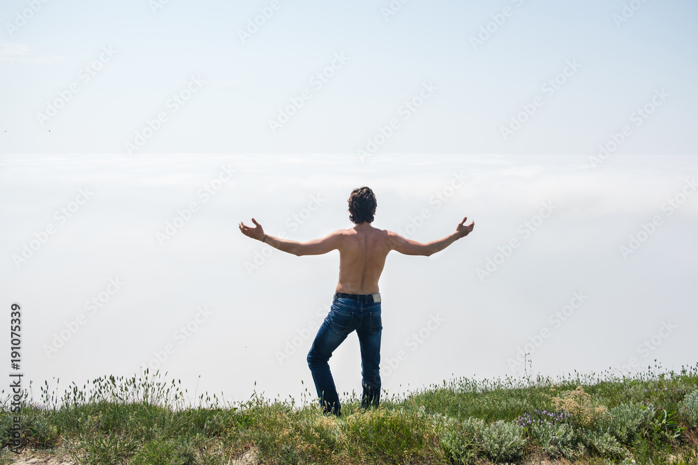 Young man with naked torso in jeans and glasses is standing on the edge of a cliff and looks at the fog. Young man with naked torso in jeans and glasses is standing on the edge of a cliff and looks at the fog.