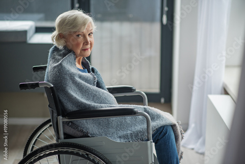Cheerless senior female sitting in invalid chair in front of the window. She is looking at camera with sad look