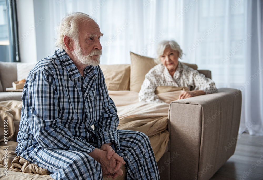 Upset old man sitting on bed with sad look. His woman sitting in bed under the blanket and looking at him with sympathy Upset old man sitting on bed with sad look. His woman sitting in bed under the blanket and looking at him with sympathy
