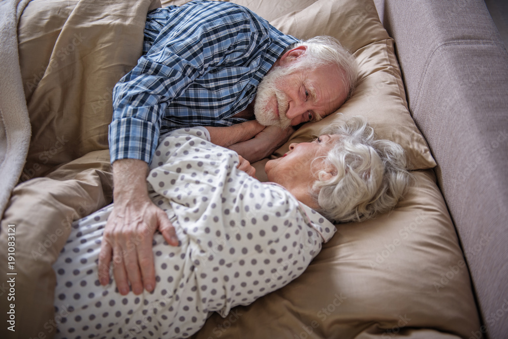 Pleased elderly couple lying in bed. Old woman and man looking at each other and smiling while embracing