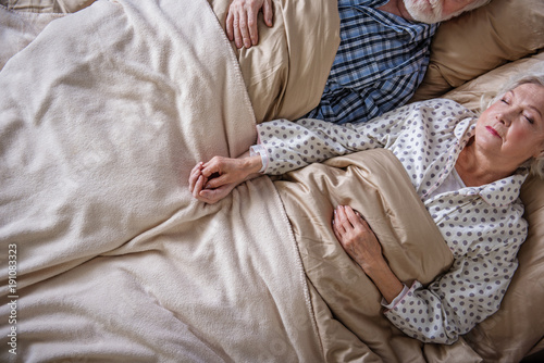 Top view of tranquil pensioners sleeping in bedroom. They are holding hands of each other