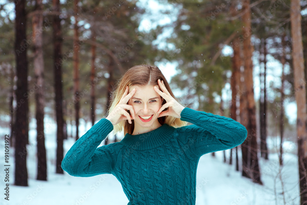 Young beautiful girl outdoors in winter. Attractive girl looking at camera smiling and holding hands near face, winter background is snowing. Portrait, close-up, space for text.