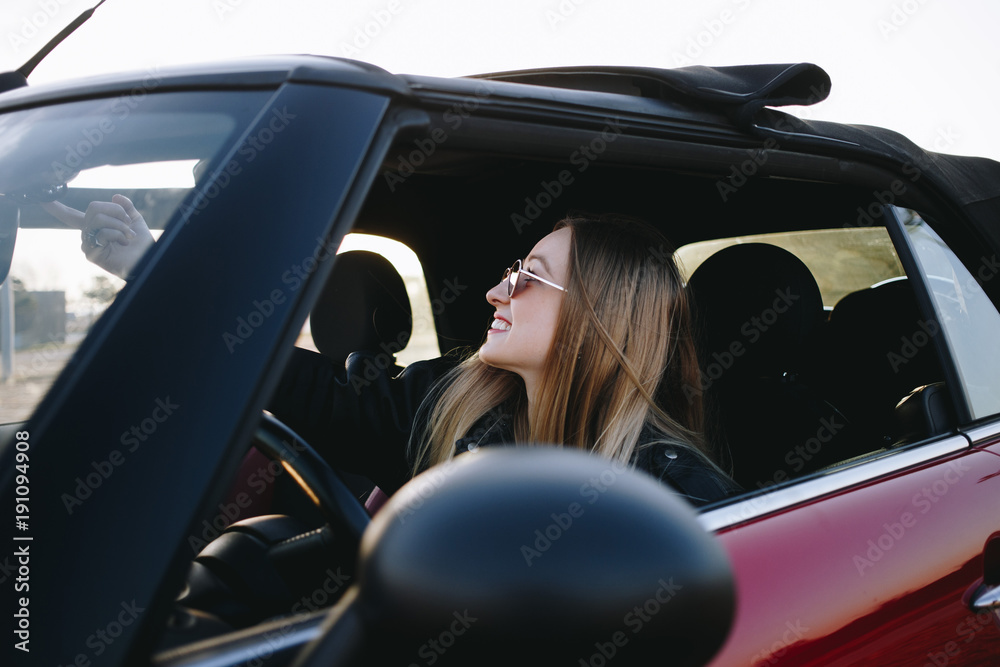 Beautiful and hip young blonde woman or teenager, hipster or millennial sits inside convertible car and opens electronical button roof top of cabriolet. First vehicle and driving license practice