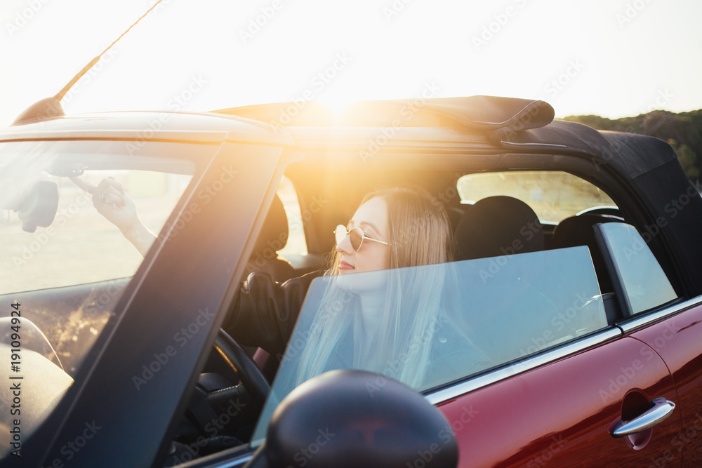 Pretty and good looking young blonde woman or model, sits inside convertible cabriolet car, before opening roof, smiles during beautiful sunset. proud new car owner teenager dream come true