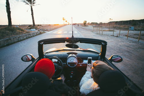 Wide angle shot of couple of teenagers or fashion internet influencers drive in cabriolet convertible mini car into sunset on palm alley. Teenage dream, forever young and happiness concept in summer