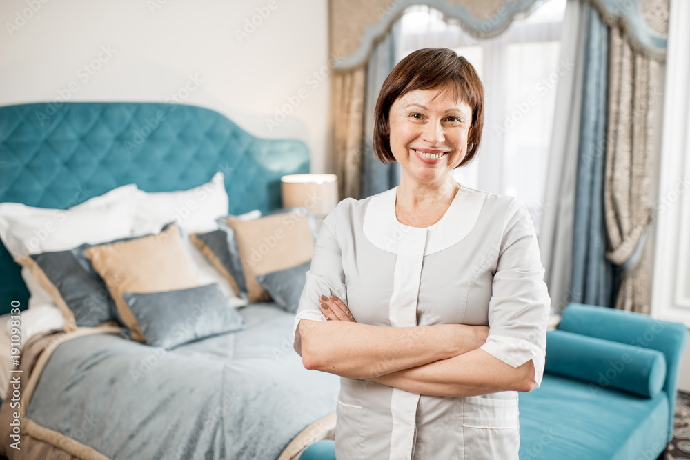 Portrait of a senior chambermaid in uniform standing in the clean hotel bedroom Portrait of a senior chambermaid in uniform standing in the clean hotel bedroom