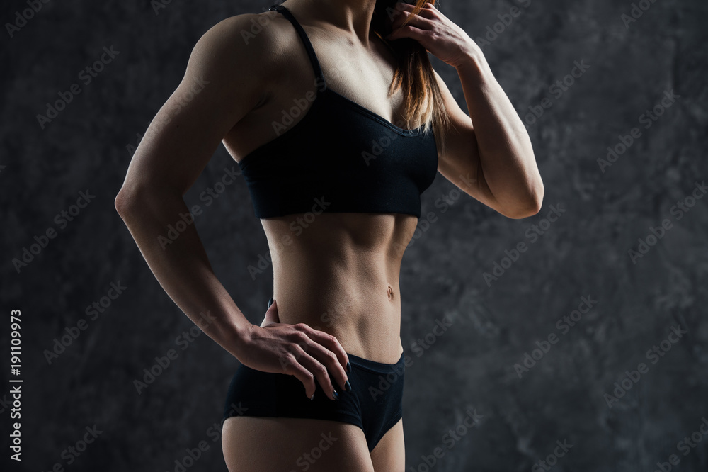 Obraz premium Side view of muscular fitness female model standing on black background. Young woman wearing sports bra looking down in thought.
