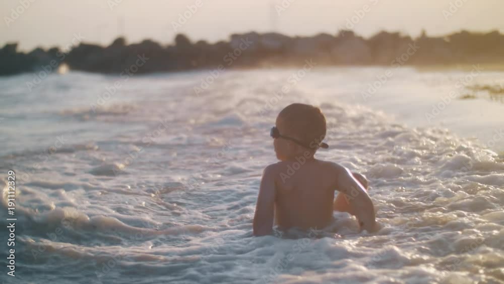 Slow motion shot of a boy lying in shallow sea water and waves hit him with splashes when rolling in on the shore, shot at sunset. Enjoyable vacation time Slow motion shot of a boy lying in shallow sea water and waves hit him with splashes when rolling in on the shore, shot at sunset. Enjoyable vacation time