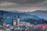 Elevated view of Banska Bystrica old town at sunset with colorful houses, snowy Krizna peak in Velka Fatra range, towers of Mestsky Hrad barbican and Nanebovzatia Panny Marie church, Slovakia Europe