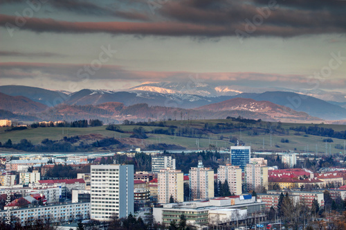 Row of socialist era tower blocks and houses in Banska Bystrica outskirts with snow-capped sunlit Velka Chochula peak of Nizke Tatry mountain range at winter sunset from Urpin hill Slovakia Europe