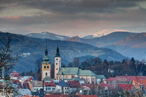 Elevated view of Banska Bystrica old town at sunset with colorful houses, snowy Krizna peak in Velka Fatra range, towers of Mestsky Hrad barbican and Nanebovzatia Panny Marie church, Slovakia Europe © nogreenabove2k