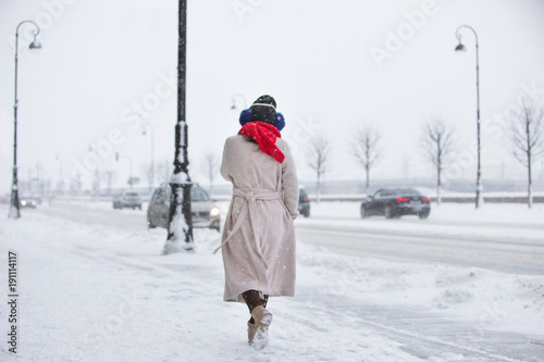 Female in beige coat, red scarf  walking on empty street during snowfall/ heavy snowfall and winter time in St. Petersburg/ Brightly dressed person in winter gloomy time/ City lifestyle concept