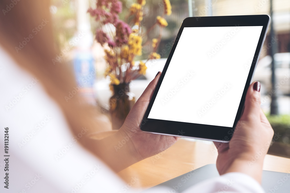 Mockup image of woman's hands holding black tablet pc with blank white desktop screen , laptop and coffee cup on wooden table in cafe