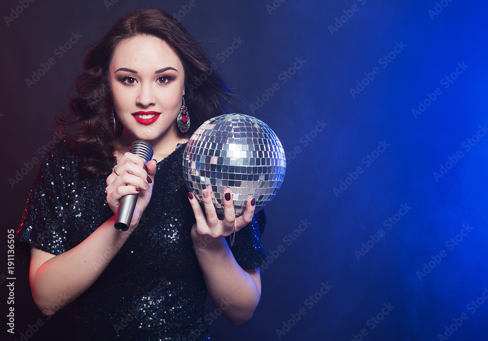 Naklejka premium lifestyle and people concept: woman wearing black dress, holding disco ball and singing into microphone
