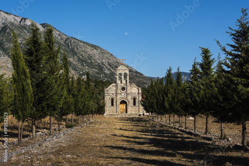 Small idyllic stone church flanked by coniferous trees in warm evening light with moon on the sky in front of a mountain range near Theth, northern Albania