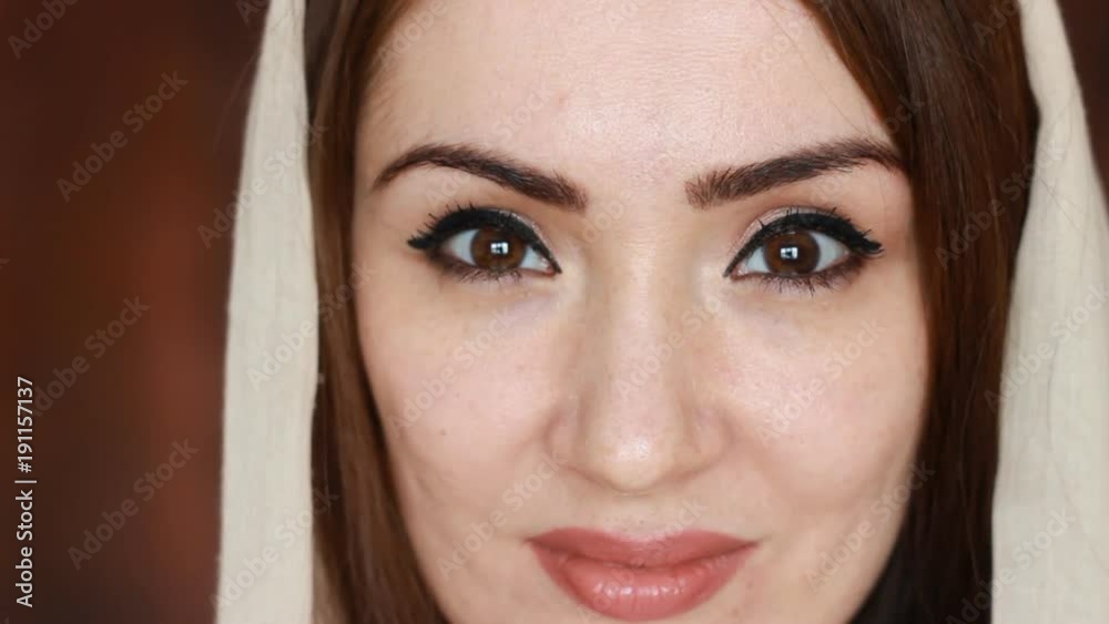 Close-up portrait of young and mysterious an arab woman in a light headscarf, called a hijab,smiles and looks at the camera. Beautiful muslim girl on a dark brown background. Close-up portrait of young and mysterious an arab woman in a light headscarf, called a hijab,smiles and looks at the camera. Beautiful muslim girl on a dark brown background.