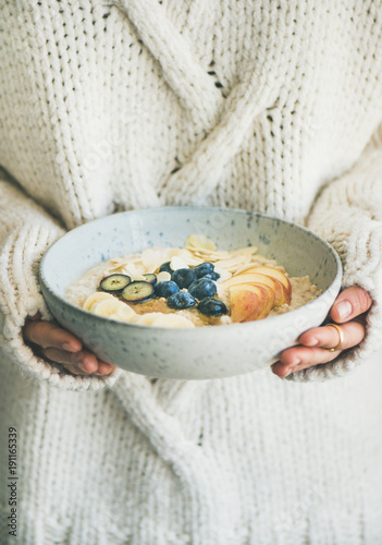 Healthy winter breakfast in bed. Woman in woolen sweater holding bowl of vegan almond milk oatmeal porridge with berries, fruit and almonds. Clean eating, vegetarian food concept