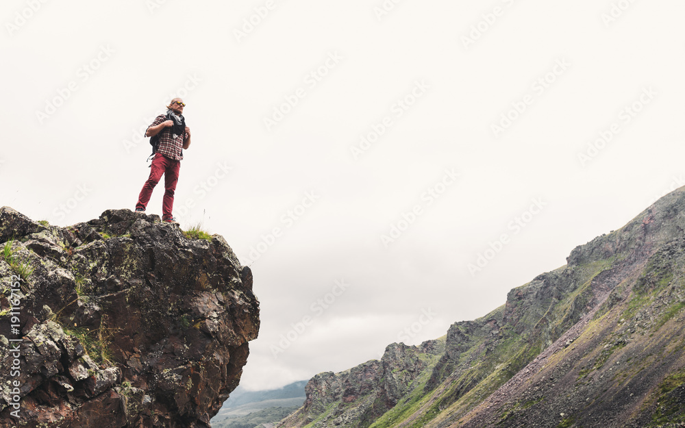 Young Man With Backpack Standing On Rocky Stone In Summer Mountains And Looking Into The Distance. Wanderlust Travel Journey Concept