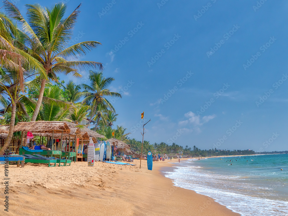 Strandszenerie am Hikkaduwa Beach mit vielen Kokospalmen, feinem Sandstrand und Palmblatt-Hütten auf der bezaubernden tropischen Insel Sri Lanka im Indischen Ozean