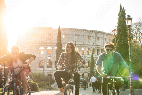 Three young friends tourists riding bikes in colle oppio park in front of colosseum on road with trees at sunset in Rome