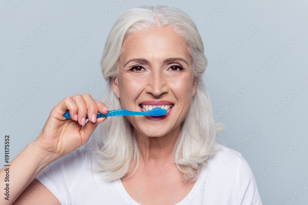 Concept of cleaning teeth is a correct way. Close up photo of happy cheerful mature old lady with good visage brushing her teeth with a blue toothbrush, isolated on gray background Concept of cleaning teeth is a correct way. Close up photo of happy cheerful mature old lady with good visage brushing her teeth with a blue toothbrush, isolated on gray background