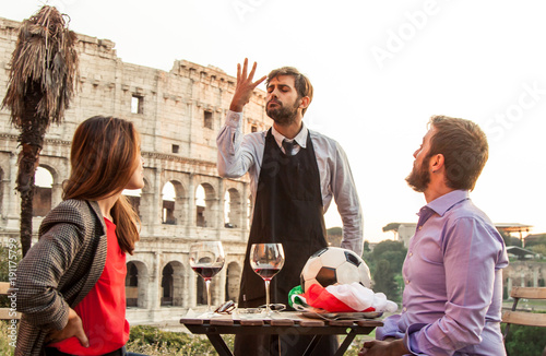 Elegant waiter serving football with italian flag to man of romantic young couple sitting at restaurant table in front of colosseum in rome at sunset. Girl is annoyed