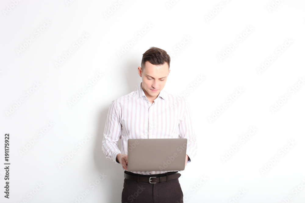 Smiling confident young man, no tie, holding grey laptop device and typing while standing against solid white wall. Wireless internet, wifi, enter password. Shaved, no facial hair. Isolated background Smiling confident young man, no tie, holding grey laptop device and typing while standing against solid white wall. Wireless internet, wifi, enter password. Shaved, no facial hair. Isolated background